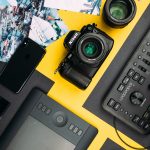 Graphic design tools (camera, computer, etc.) on a yellow desk