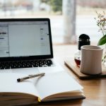 An open laptop and book on a desk, near a coffee cup and vase of flowers
